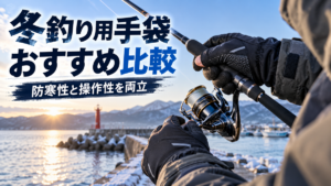 冬の海辺で防寒フィッシンググローブを着用し、スピニングリール付きの釣り竿を操作する釣り人。